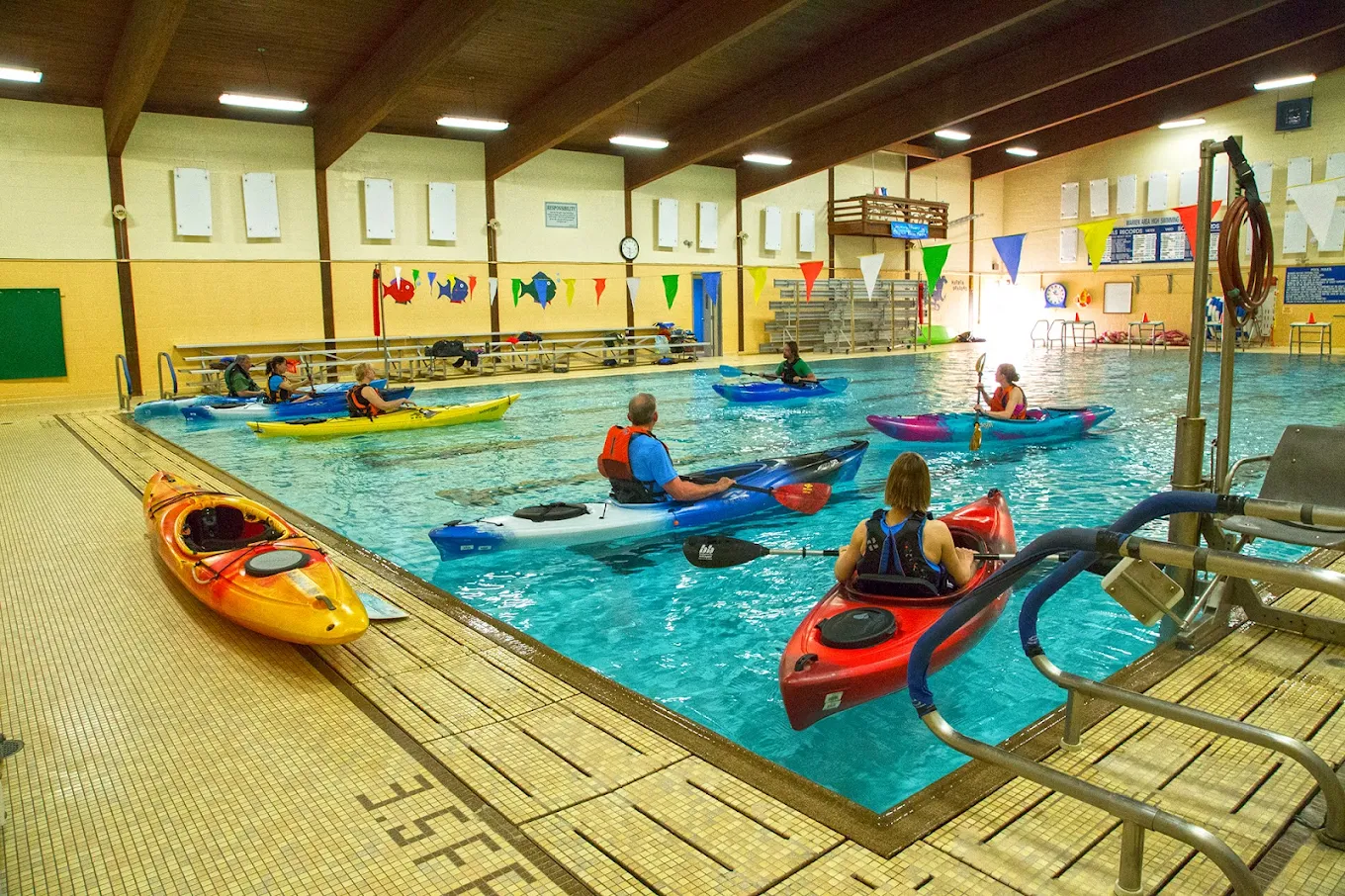 Seven people, each in their own kayak, in an indoor pool.