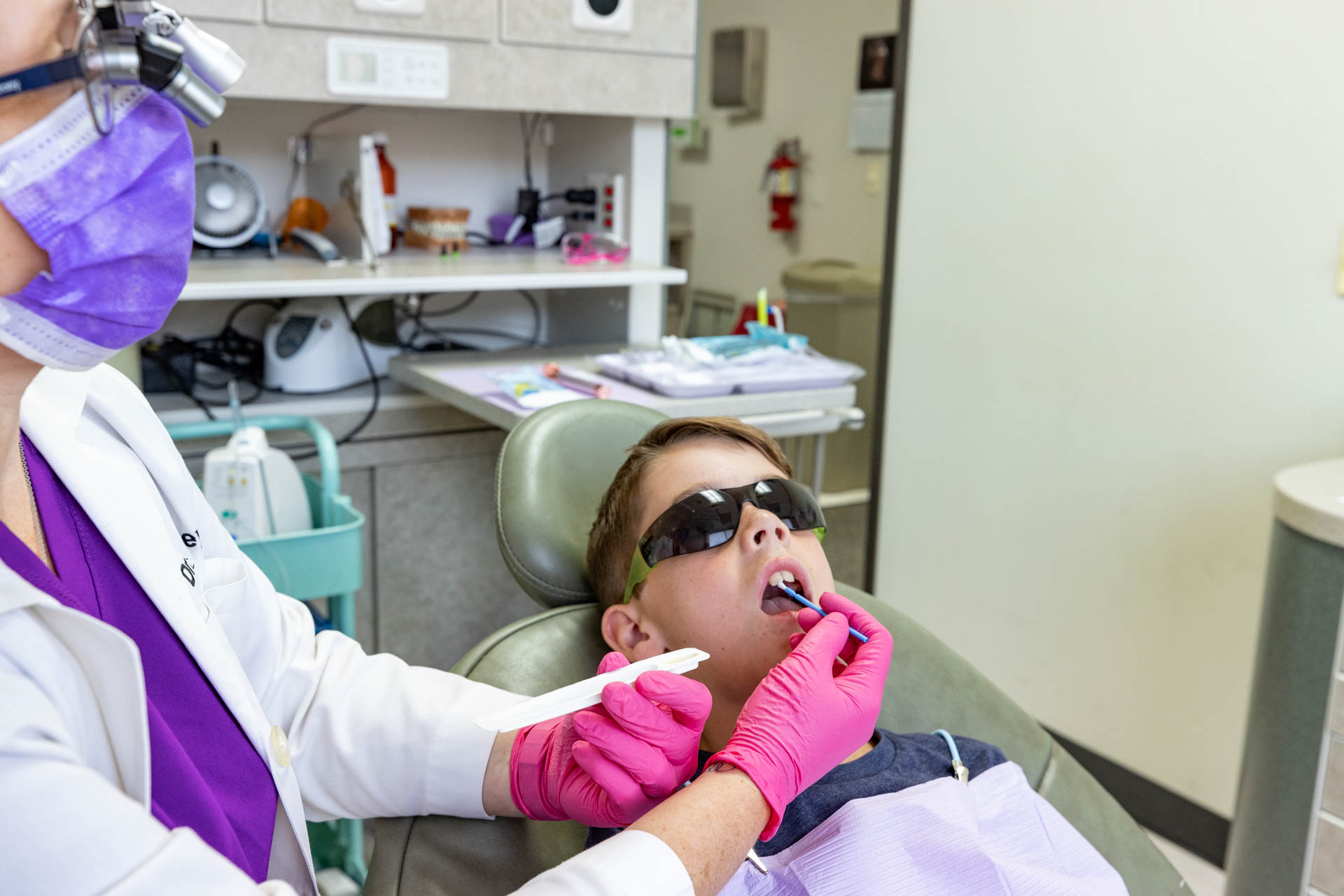 A child wearing protective eyewear laying in a dental chair while a dentist does a procedure in the child's mouth.