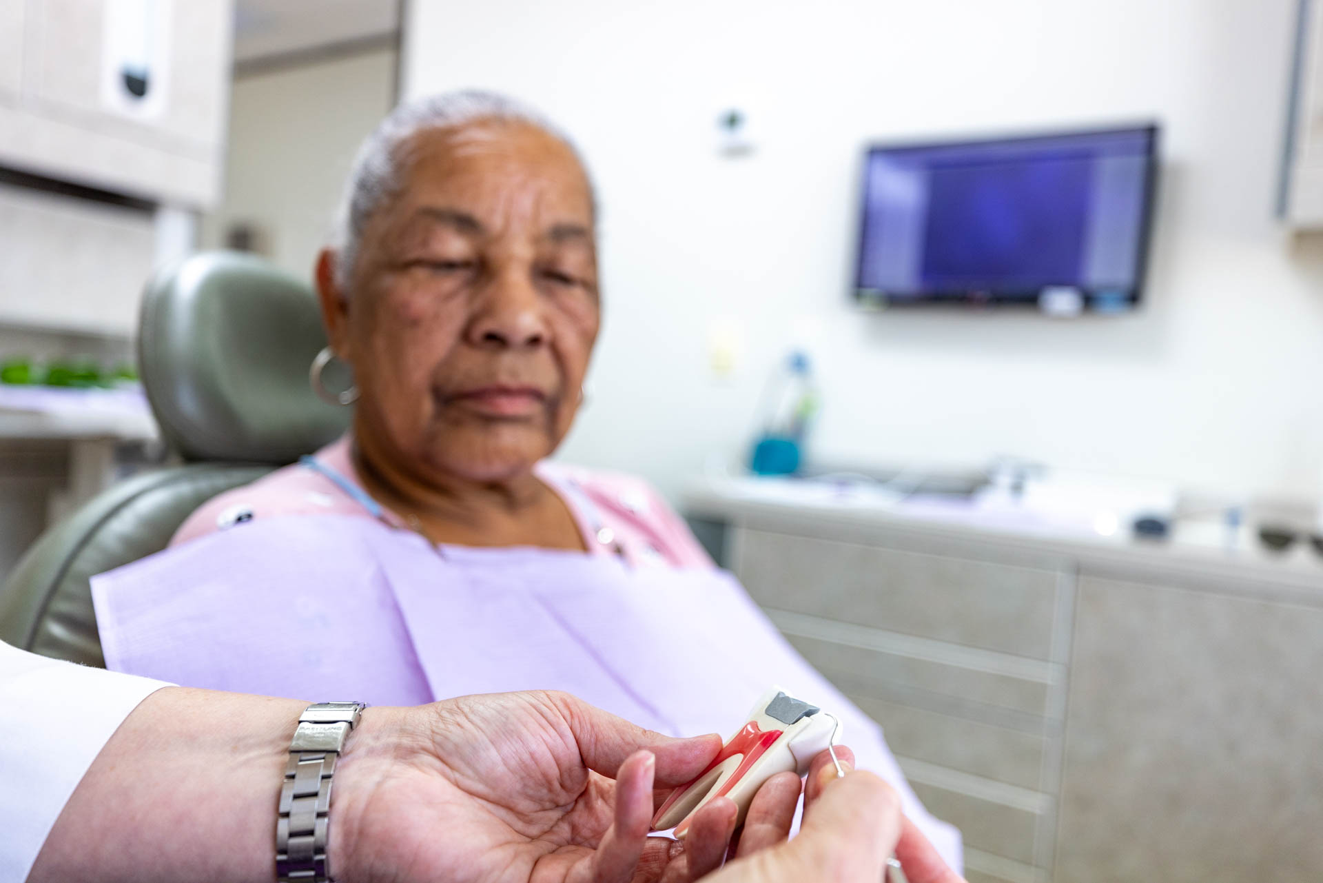 An older woman blurred in the background with hands holding a dental tool in the foreground.