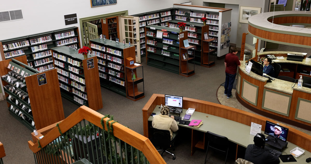 An interior high-angle view from the second-floor balcony of the Warren Public Library, looking down at a bright, open central atrium with bookshelves, seating areas, and modern geometric carpeting.