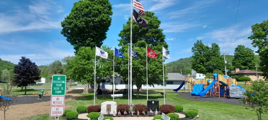 A sunny community park featuring a veterans memorial with multiple military flags flying, alongside a bright blue children's playground in the background.