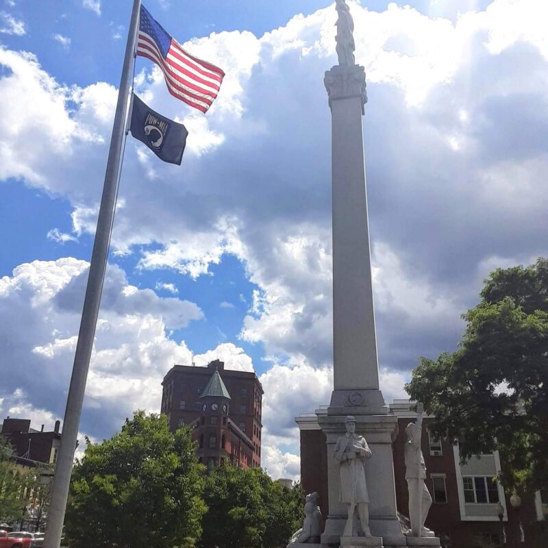 A tall stone obelisk monument with statues of soldiers at its base, standing next to a flagpole flying the American and POW/MIA flags under a bright blue sky with fluffy clouds.