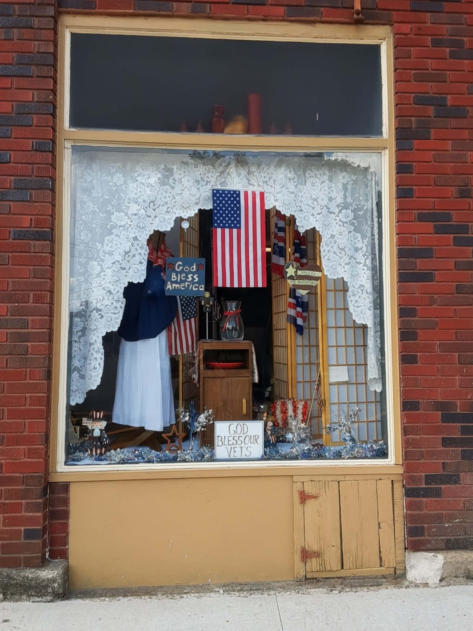 A brick storefront window display featuring an American flag, lace curtains, and signs that say "God Bless America" and "God Bless Our Vets."