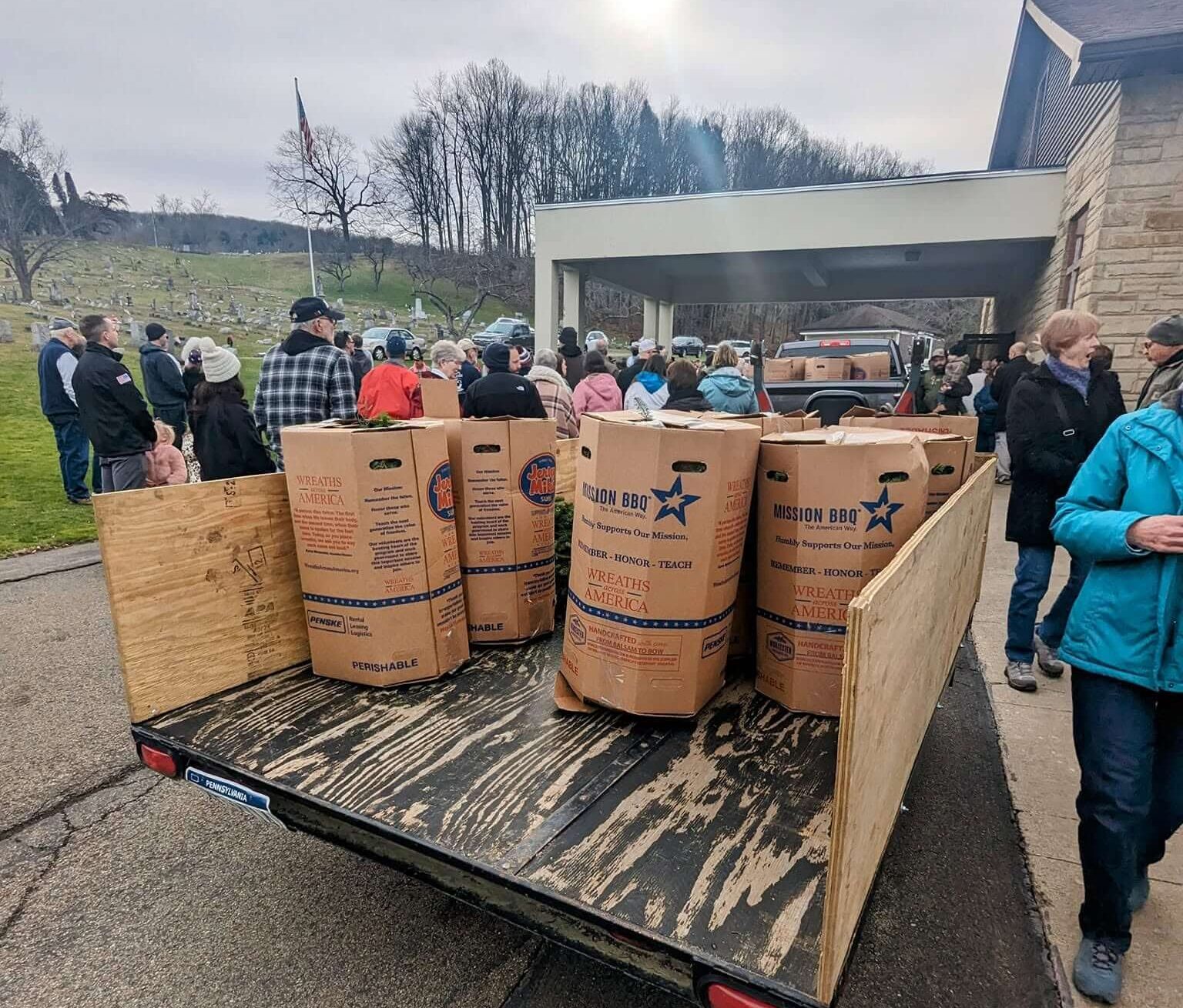 A flatbed trailer carrying Wreaths Across America boxes at a cemetery, with local volunteers gathered in the background.